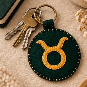 May include: A circular dark green felt keychain featuring a yellow embroidered Taurus zodiac symbol in the center. The edges are stitched with yellow thread, and it is attached to a brass-toned metal split ring holding two silver-toned keys.