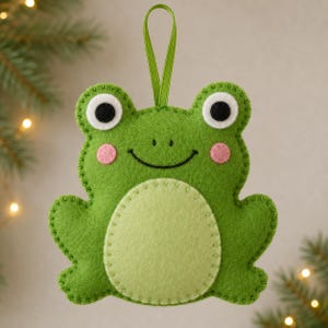 Könnte beinhalten: Ein grüner Filz-Frosch-Anhänger mit einem hellgrünen Bauch, weiß-schwarzen Augen und rosa Wangen. Der Frosch hat eine genähte Kontur und eine grüne Bandschlaufe an der Oberseite zum Aufhängen. Das Design ist flach mit einer weichen Textur.