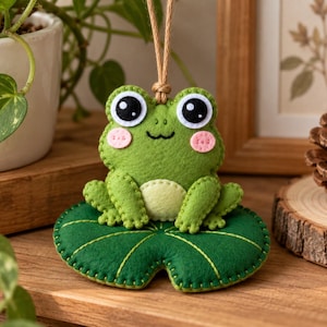 Könnte beinhalten: Ein hängendes Filzornament in Form eines grünen Frosches, der auf einem dunkelgrünen Seerosenblatt sitzt. Der Frosch hat große schwarz-weiße Augen, rosa Wangen und einen hellgrünen Bauch. Er ist an einer dünnen beigen Schnur befestigt.