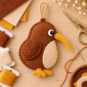 Può includere: Un ornamento in feltro a forma di kiwi marrone con pancia color crema, un lungo becco giallo e piedi gialli. L'uccello presenta un piccolo occhiello sulla parte superiore per appenderlo ed è realizzato con cuciture marroni visibili sui bordi.