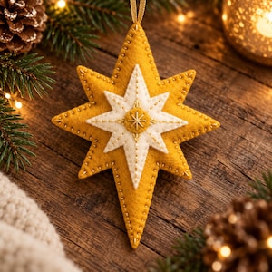 May include: A star-shaped felt ornament featuring a large mustard yellow base layer topped with a smaller white felt star. The design includes gold-colored decorative stitching along the edges and a central circular accent with a stitched star pattern.