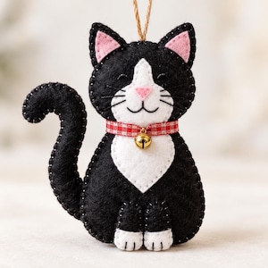 May include: A black and white felt cat ornament with a red and white gingham collar and a small gold bell. The cat has pink inner ears, stitched facial features, and a curved tail, suspended by a thin brown string for hanging display.