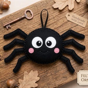 May include: A black felt spider ornament with white eyes, pink cheeks, and a smiling mouth. The spider is on a wooden surface with keys, acorns, and a tag that says "Handmade with Love". The tag also says "Felt Spider Ornament".