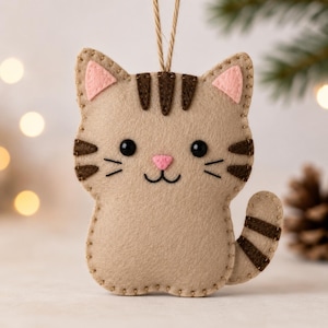 Può includere: Un ornamento in feltro color marrone chiaro a forma di gatto con strisce marrone scuro su fronte, guance e coda. Il gatto presenta orecchie interne in feltro rosa, un naso rosa, occhi a perla neri e una bocca ricamata con baffi e cordino.