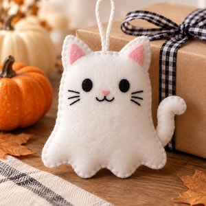 May include: A white felt cat ornament with a ghost-like body shape, featuring pink inner ears, a small pink nose, black circular eyes, and stitched black whiskers. The ornament includes a white cord loop at the top for hanging purposes.
