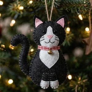 Puede incluir: Adorno navideño de gato en blanco y negro, con orejas rosas, nariz rosa y un collar a cuadros rojos y blancos. Tiene una pequeña campana dorada y cuelga de una cuerda. Una encantadora decoración festiva.