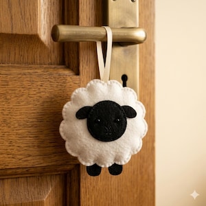 May include: A round, scalloped-edge felt ornament shaped like a sheep with a white body and a black face. The sheep features two small black ears, two tiny black bead eyes, and a stitched nose. A thin cream-colored ribbon loop is attached to the top.