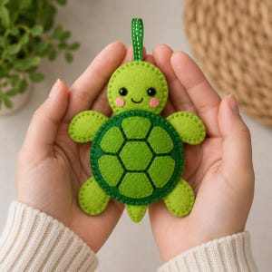 Könnte beinhalten: Ein Schildkröten-Anhänger aus Filz mit einem hellgrünen Kopf, schwarzen Augen und rosa Wangen, einem dunkelgrünen Panzer mit genähtem Sechseckmuster, vier hellgrünen Flossen und einer kleinen grünen Bandschlaufe zum Aufhängen an der Oberseite.
