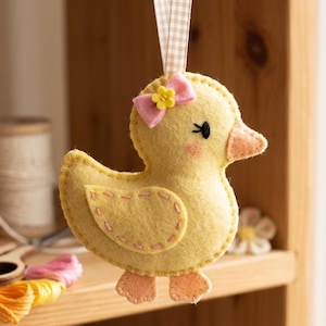 May include: A handmade yellow felt duck ornament with a pink bow and flower on its head. The duck has a peach-colored beak and feet, with stitched details. A gingham ribbon is attached for hanging.