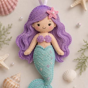 Op de afbeelding: Een handgemaakte vilten zeemeermin-ornament met paars haar, een roze zeester en een turquoise staart. De zeemeermin heeft een schelpen top en is versierd met geborduurde details. Een wit lint is bevestigd om op te hangen.