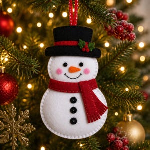 May include: A felt snowman ornament featuring a white body, three black buttons, a red scarf, and a black top hat adorned with a red ribbon and green holly leaves. The snowman has pink cheeks and an orange carrot nose, suspended by a red ribbon.