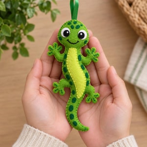 May include: A felt lizard ornament in shades of bright green and lime green with dark green spots. The lizard features large white and black eyes, a stitched smile, and a green ribbon loop at the top for hanging. The body is crafted from felt.