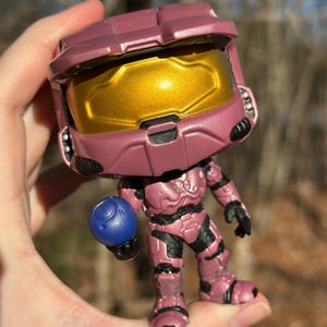 以下が含まれることがあります： Haloのビデオゲームシリーズのキャラクターのピンク色のアクションフィギュア。フィギュアは金色のバイザー、黒のアクセントがあり、青いオーブを持っています。フィギュアの高さは約10cmです。