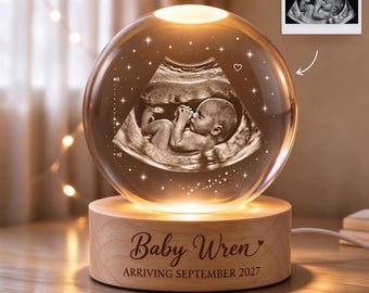 Peut inclure: Une lampe sphérique en verre transparent présentant une image échographique gravée au laser en 3D d'un fœtus à l'intérieur. La sphère repose sur une base en bois ronde gravée du texte Baby Wren et ARRIVING SEPTEMBER 2027, éclairée par le bas.