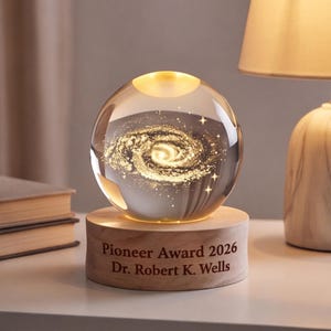 Op de afbeelding: Een heldere glazen bol met een intern lasergegraveerd gouden spiraalstelselontwerp, rustend op een cilindrische lichtgekleurde houten voet. De voet is gegraveerd met de tekst: Pioneer Award 2026, Dr. Robert K. Wells, in donkere letters.