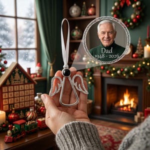 May include: A silver-toned angel ornament featuring a circular black center, crystal-encrusted wings, and a white ribbon loop. A circular inset displays a portrait of an older man with the text Dad 1948 - 2026, framed by a decorative border.