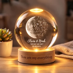 Könnte beinhalten: Eine klare Glaskugellampe mit einem internen, lasergravierten Mond- und Sternbildmotiv. Die Kugel zeigt den Text Olivia & Noah, THE DAY WE MET und 09.08.2025. Sie steht auf einem runden Holzsockel mit der Gravur You are my everything.
