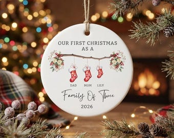 以下が含まれることがあります： 紐で吊るされた丸い白い陶器のクリスマスオーナメント。"OUR FIRST CHRISTMAS AS A"という文字の下に、"DAD"、"MOM"、"LILY"とラベル付けされた3つの赤い靴下が描かれた枝のイラストがあり、その下に"Family Of Three"と"2026"が記されています。
