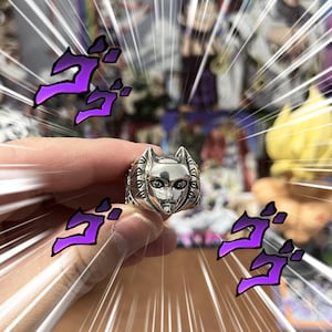 Op de afbeelding: Een zilverkleurige metalen ring met een gebeeldhouwd, gestileerd katachtig gezicht met puntige oren en gedetailleerde gelaatstrekken. De ring heeft een gepolijste afwerking en wordt geaccentueerd door paarse Japanse katakana-tekens op de voorgrond.