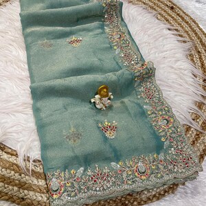 May include: A seafoam green saree with detailed embroidery along the edges and across the fabric. The embroidery includes red, yellow, and silver details. A small decorative figurine is placed on the saree.
