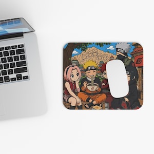 Puede incluir: Alfombrilla de ratón rectangular con una colorida ilustración de estilo anime de varios personajes. El diseño incluye un fondo de montaña, edificios y personajes comiendo ramen. Un ratón de ordenador blanco descansa sobre la alfombrilla.