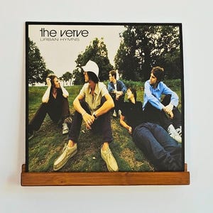 Op de afbeelding: Een vierkante vinyl platenhoes voor het album Urban Hymns van The Verve, met vier personen zittend op een grasheuvel. De hoes toont de bandleden in vrijetijdskleding, inclusief een witte vissershoed, tegen een achtergrond van bomen.