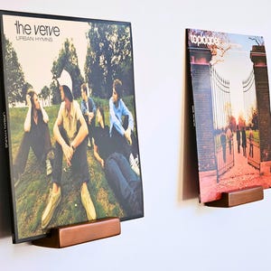 Op de afbeelding: Twee vinylplaten tentoongesteld op kleine houten wandplanken. De linker albumhoes toont The Verve Urban Hymns met een groep mensen in het gras. De rechter albumhoes toont Toploader met een poortingang bij zonsondergang.