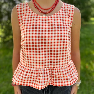 May include: A sleeveless top featuring a red and white gingham check pattern with a round neckline and a gathered peplum hem. The garment is paired with a layered red beaded necklace that includes a small silver-toned star-shaped pendant.
