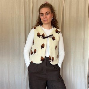 May include: A cream-coloured fleece waistcoat featuring a repeating pattern of brown acorns. The sleeveless garment has a round neckline and a dark brown ribbon tie closure at the centre front. It is worn over a plain white long-sleeve shirt and dark trousers.