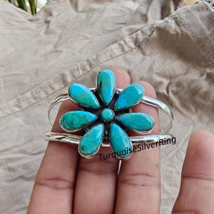 May include: A silver cuff bracelet featuring a turquoise flower design. The flower has six turquoise petals surrounding a smaller turquoise center. The bracelet is held in a hand, with the text "TurquoiseSilverRing" visible.