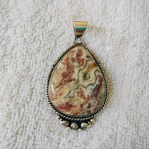 May include: A teardrop-shaped pendant featuring a polished stone with swirling patterns of cream, tan, and deep reddish-brown. The stone is set in a silver-toned metal frame with a twisted rope border and a decorative beaded accent at the base.