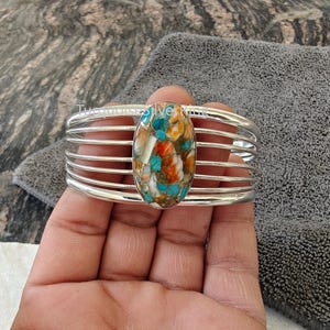 Könnte beinhalten: Ein silbernes Armband mit einer zentralen ovalen Einlage aus mehrfarbigem Stein, einschließlich Türkis-, Orange- und Weißtönen, eingefasst in einen polierten Metallrahmen, der aus fünf parallelen Bändern besteht, die das Handgelenk umschließen.