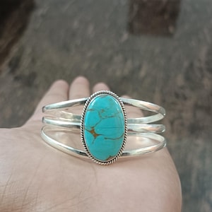 May include: A silver cuff bracelet featuring a central oval turquoise stone with brown matrix veining. The stone is set within a twisted rope border and mounted on a triple-band silver frame that splits into three distinct polished metal segments.