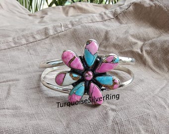 May include: A silver cuff bracelet with a large flower design. The flower has pink, turquoise, and white petals with a bronze center. The text "TurquoiseSilverRing" is below the bracelet.