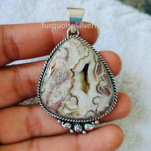 May include: A teardrop-shaped pendant featuring a polished Crazy Lace Agate stone with swirling white, cream, and reddish-brown patterns and a central geode cavity. The stone is set in a twisted silver frame with decorative metal bead accents.