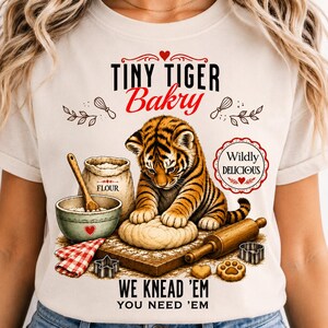 May include: A cream-coloured t-shirt featuring a graphic of a tiger cub kneading dough on a wooden board. The design includes a flour bag, a mixing bowl, a rolling pin, cookie cutters, and the text "TINY TIGER Bakry", "WILDLY DELICIOUS", "WE KNEAD 'EM", and "YOU NEED 'EM".