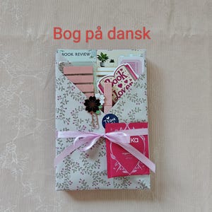 May include: A gift-wrapped package with a floral pattern, tied with a pink ribbon. The package contains a variety of items, including a small plant, stickers, and a bookmark. A black flower clip and a "Thank You" sticker are also visible.