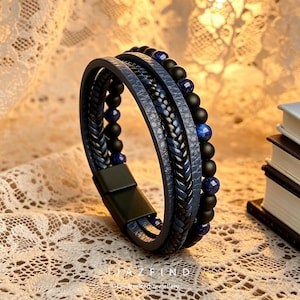May include: A multi-strand bracelet featuring blue leather straps, braided accents, and black and blue beads. The bracelet has a magnetic clasp and is displayed on a textured surface. The bracelet is a piece of handcrafted jewelry.