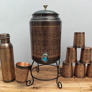 Puede incluir: Un juego de dispensador de agua de color cobre con una botella de agua, una taza y seis tazas pequeñas a juego. El dispensador tiene un grifo y se apoya en un soporte de metal negro. Todos los artículos presentan un patrón ornamental repetido.