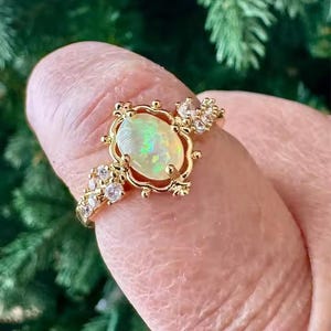 Peut inclure: Une bague en or avec une opale ovale centrale aux reflets irisés verts et orange. La monture est ornée de petites pierres claires taillées en rond, incrustées le long de l'anneau et encadrant l'opale centrale dans un support en métal décoratif.