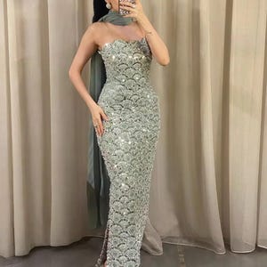 Könnte beinhalten: Ein trägerloses, bodenlanges Kleid in Meeresgrün, verziert mit aufwendigen Pailletten. Das Kleid hat einen wellenförmigen Ausschnitt und einen hohen Seitenschlitz. Ein passender Schal liegt über den Schultern. Das Model trägt silberne Sandalen.