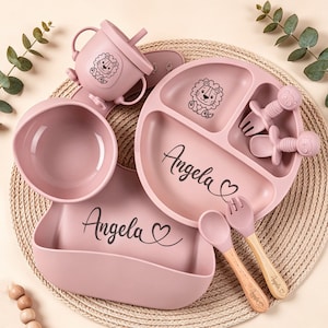 May include: A pink baby feeding set with a lion graphic and the name "Angela" in cursive. The set includes a divided plate, bowl, cup, bib, spoon, and fork. The spoon and fork have wooden handles. The set is arranged on a woven mat.