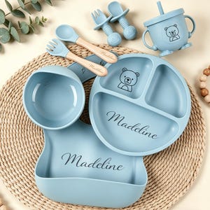 May include: A baby feeding set in light blue, featuring a divided plate, bowl, bib, cup, spoons, and forks. The plate and cup have a bear graphic. The name "Madeline" is printed on the plate and bib.