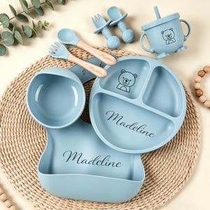 May include: A baby feeding set in light blue, featuring a divided plate, bowl, bib, cup, spoons, and forks. The plate and cup have a bear graphic. The name "Madeline" is printed on the plate and bib.