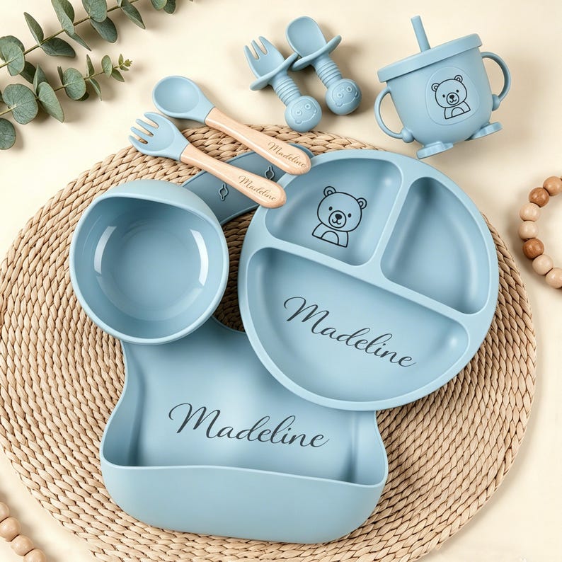 May include: A baby feeding set in light blue, featuring a divided plate, bowl, bib, cup, spoons, and forks. The plate and cup have a bear graphic. The name "Madeline" is printed on the plate and bib.