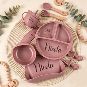 May include: A set of baby feeding essentials in a dusty rose color. The set includes a divided plate, bowl, bib, sippy cup, spoon, and fork. Each item is personalized with the name "Nicola" and a bow design. The spoon and fork have wooden handles.