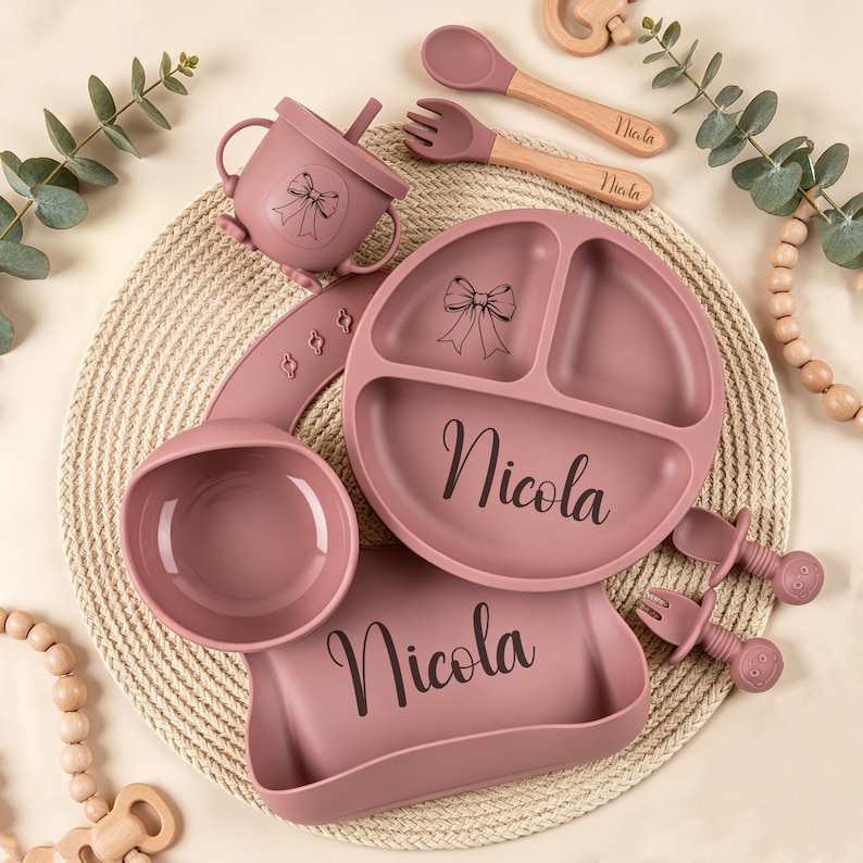 May include: A set of baby feeding essentials in a dusty rose color. The set includes a divided plate, bowl, bib, sippy cup, spoon, and fork. Each item is personalized with the name "Nicola" and a bow design. The spoon and fork have wooden handles.