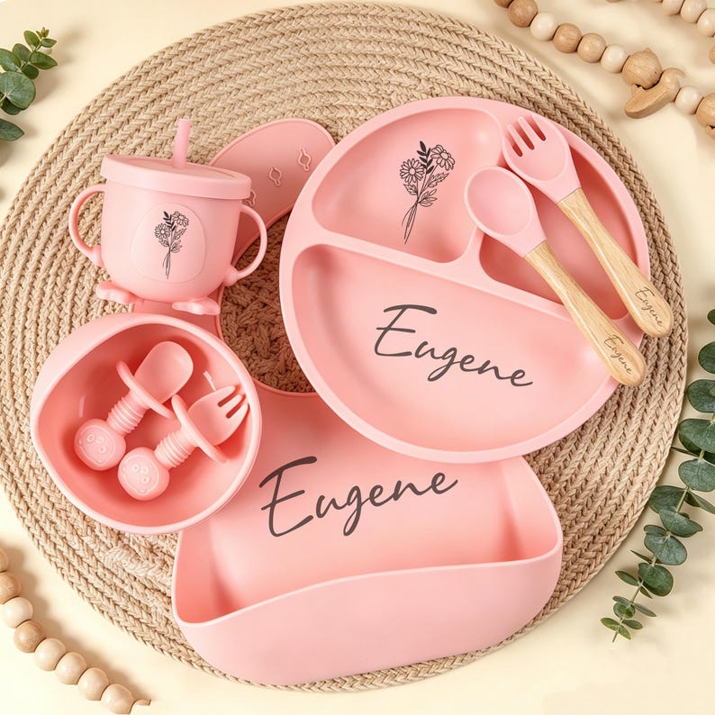May include: A pink baby feeding set featuring a divided plate, bowl, cup with straw, bib, and utensils. The set includes a plate, bowl, and bib with the name "Eugene" printed on them. The utensils have wooden handles. Floral designs are also present.