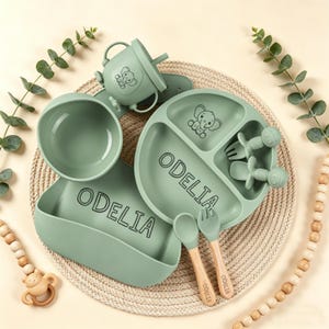 May include: A sage green baby feeding set with the name "ODELIA" printed on the bib and plate. The set includes a divided plate, bowl, cup, bib, spoon, fork, and teether. Each piece has a cartoon elephant design.