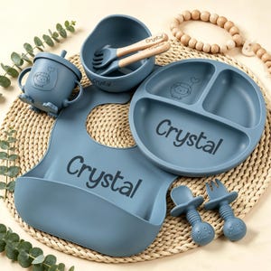 May include: A blue baby feeding set includes a divided plate, bowl, bib, cup with straw, and two utensils. The set features a sheep graphic and the name "Crystal". The bowl and utensils have wooden handles.